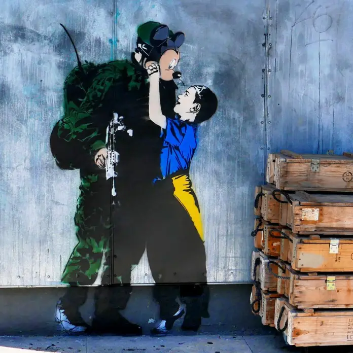 You never know what’s behind the mask by graffit artist Hijack. Shows a child in Ukrainian colours taking a micky mouse mask off a Russian soldier.