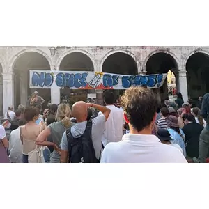A crowd of protesters gathers in front of a historic Venetian building with arched stone columns, holding a large hand-painted banner that reads “NO SPACE FOR BEZOS!” accompanied by a cartoon rocket. The mood is lively and defiant, with people standing shoulder to shoulder, some filming or raising their fists.