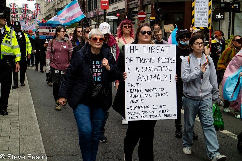 A group of protesters march down a city street holding trans pride flags and placards. At the centre, a person holds a large sign reading: “THE THREAT OF TRANS PEOPLE IS A STATISTICAL ANOMALY! FACT: EHRC WANTS TO HUMILIATE TRANS PEOPLE. SUPREME COURT RULING DID NOT HEAR FROM ONE TRANS PERSON.” The crowd includes people of various ages and genders, with police officers visible to the left and Union Jack flags hanging in the background. The mood is serious but determined.