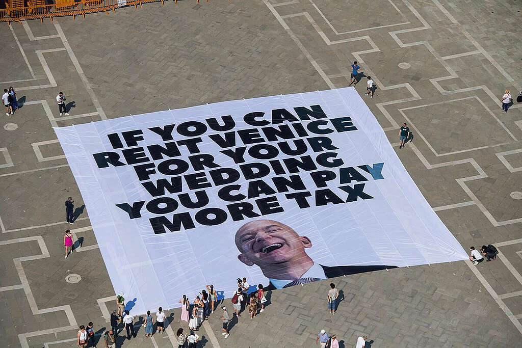 A large protest banner laid out in a Venetian square reads “IF YOU CAN RENT VENICE FOR YOUR WEDDING YOU CAN PAY MORE TAX” in bold black letters, featuring a smiling image of Jeff Bezos. Tourists and passers-by gather around the banner, taking photos and observing the scene.
