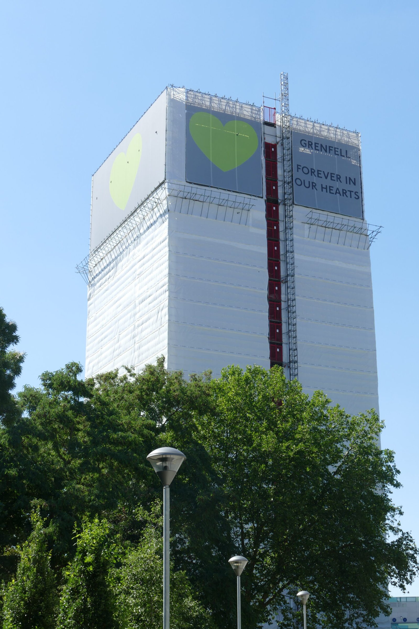 Grenfell Tower covered in white sheeting with large green heart symbols and the words "GRENFELL FOREVER IN OUR HEARTS" displayed at the top. A red construction lift runs vertically up the centre, with trees and lampposts in the foreground under a clear blue sky.