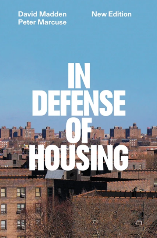 Book cover of In Defense of Housing by David Madden and Peter Marcuse. The image shows a dense urban skyline of low-rise and high-rise apartment buildings under a clear blue sky. The title is prominently displayed in large, bold white capital letters at the centre, partially obscuring the skyline. The authors’ names appear in smaller white text at the top corners, with “New Edition” noted in the top right. The overall design conveys a serious and grounded tone, centred on the urban housing landscape.