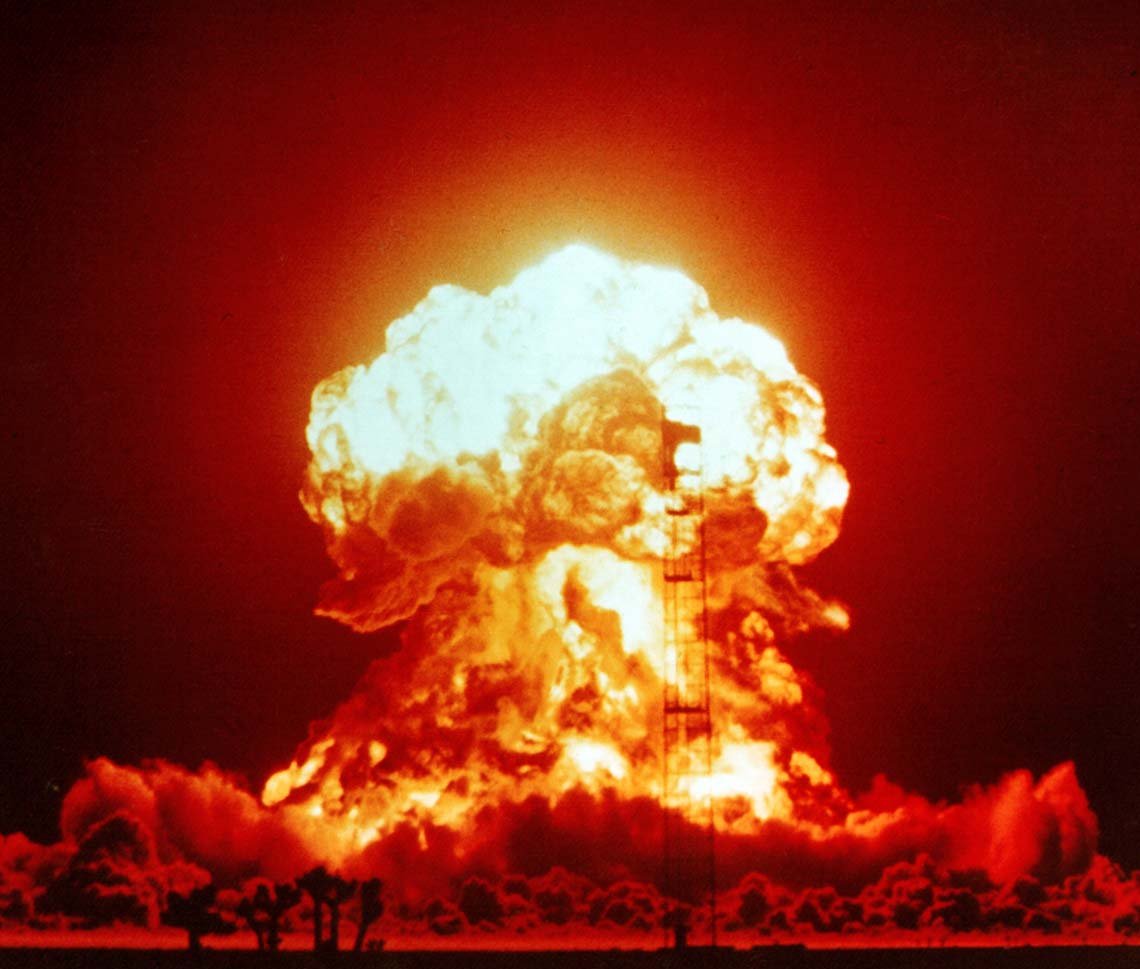 A vivid red and orange photograph of a nuclear explosion during Operation Upshot-Knothole. A massive fireball and mushroom cloud dominate the image, illuminating the night sky. A steel test tower is visible in front of the blast, and silhouetted Joshua trees and human figures can be seen in the foreground, emphasising the scale and intensity of the detonation.