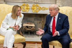 Giorgia Meloni, dressed in a white suit, leans forward intently as she listens to Donald Trump, who is seated opposite her in a navy blue suit and red tie, gesturing mid-sentence. The two sit in ornate yellow chairs in front of a fireplace adorned with elaborate gold decorations, suggesting a formal diplomatic setting.