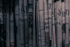 A close-up image of tightly rolled newspapers stacked vertically, with dim, moody lighting and a grainy texture that gives the scene a vintage, noir atmosphere. Some headlines and columns are partially visible, adding to the sense of layered, obscured information.