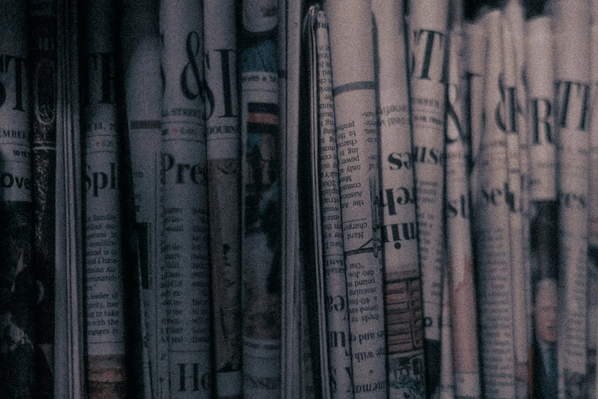 A close-up image of tightly rolled newspapers stacked vertically, with dim, moody lighting and a grainy texture that gives the scene a vintage, noir atmosphere. Some headlines and columns are partially visible, adding to the sense of layered, obscured information.