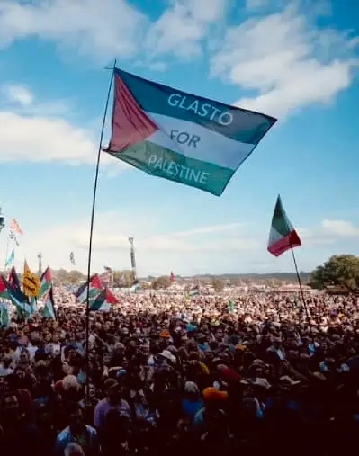A large crowd at Glastonbury Festival waves numerous Palestinian flags under a partly cloudy blue sky. Dominating the image is a prominently raised flag in the foreground reading “GLASTO FOR PALESTINE” in white letters across the Palestinian tricolour (black, white, and green horizontal stripes with a red triangle on the hoist side). The atmosphere is festive yet political, with a visible sense of solidarity.