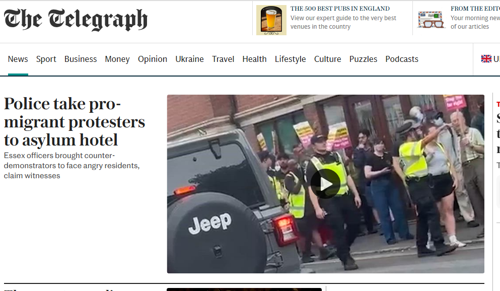 A screenshot of The Telegraph’s website front page. The main headline reads: “Police take pro-migrant protesters to asylum hotel.” The subheading says, “Essex officers brought counter-demonstrators to face angry residents, claim witnesses.” Beside the text is an image of uniformed police officers in high-visibility jackets standing in front of a group of pro-migrant protesters holding “Stand Up to Racism” placards. A black Jeep with a visible rear wheel is parked in the foreground. The scene takes place outside the Bell Hotel in Clacton-on-Sea. The mood is tense, with the police forming a line between protesters and onlookers.