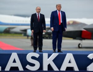 Vladimir Putin and Donald Trump stand side by side on a stage with a large “ALASKA” sign in front of them. Both wear dark suits and red ties, standing stiffly against the backdrop of aircraft, including Air Force One. A red carpet stretches out behind them, suggesting a formal state visit setting.