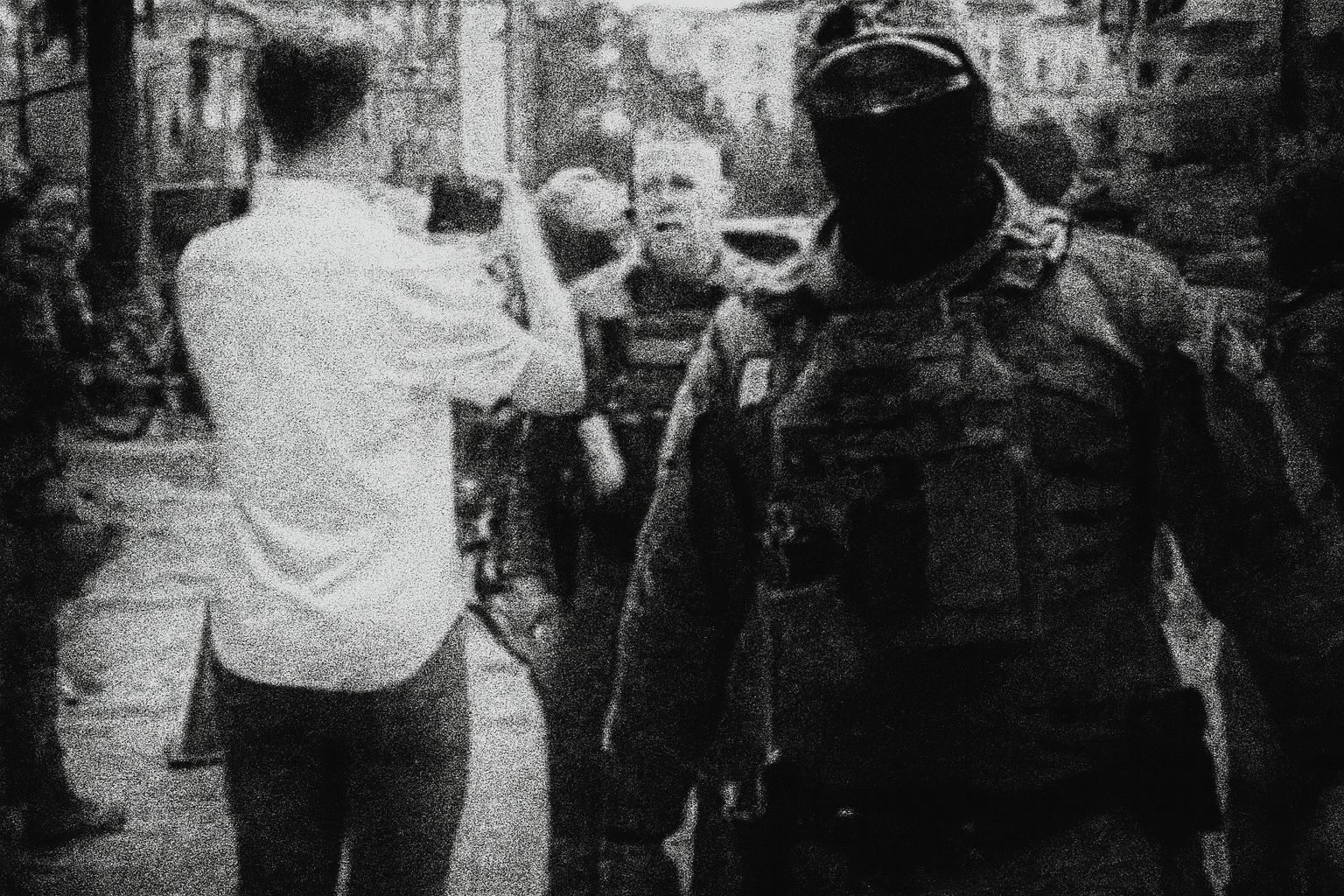 Grainy black-and-white image showing a masked, heavily armed ICE officer in tactical gear dominating the foreground, with other armed officers and a man in a white shirt filming them in the blurred background, creating a tense, menacing atmosphere.