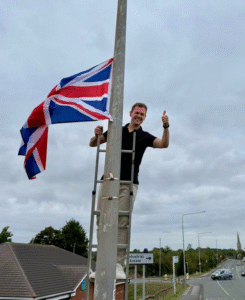 Robert Jenrick stands on a ladder attached to a lamppost, giving a thumbs-up while raising a Union Jack flag. The background shows a cloudy sky, rooftops, road signs, and a quiet road stretching into the distance.