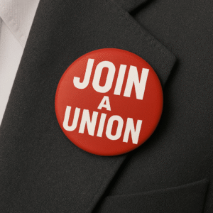 A close-up photograph of a dark grey suit jacket with a red circular badge pinned to the left lapel. The badge features bold white text that reads "JOIN A UNION" in all capital letters. The image is softly lit, with the badge clearly in focus and the texture of the suit fabric visible in the background.